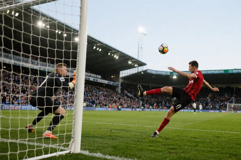 一名足球运动员在中场位置起脚吊射，足球飞跃守门员冲向球门，观众屏息凝视